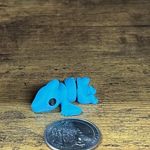 May include: A small, light blue 3D-printed lizard figurine with a black eye, lying next to a quarter coin. The lizard has a detailed, textured surface. The figurine is on a wooden surface.