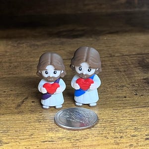 May include: Two small figurines of Jesus holding red hearts, with brown hair and white robes. A silver coin is next to the figurines. The figurines are on a wooden surface.