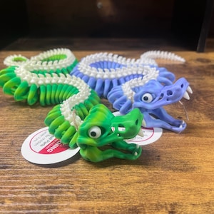 May include: Three articulated dragon toys in green, blue, and purple. The green dragon has a white spine, the blue dragon has a white spine, and the purple dragon has a white spine. Each dragon has a detailed head and body. The toys are on a wooden surface.