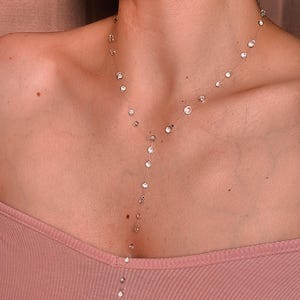 May include: A delicate lariat-style necklace featuring a thin, transparent cord adorned with evenly spaced, round, clear faceted crystals. The design includes a central vertical drop section that extends downward from the main neckline chain.