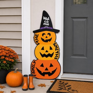 May include: A vertical wooden porch sign featuring three stacked orange jack-o'-lanterns topped with a black witch hat. The hat displays the white text "Welcome" and the pumpkins feature the black text "The Smiths Family" along the sides.