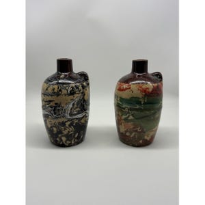 May include: Two decorative ceramic jugs with dark brown necks and handles. One jug has a marbled pattern in black, tan, and white. The other jug features a mix of red, green, and tan colors. Both jugs have a rustic, handcrafted appearance.