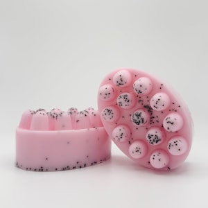 May include: Two pink soaps with black specks. One is oval-shaped with raised, round bumps. The other is rectangular with a ridged top. Both soaps have a light pink color and a speckled appearance.
