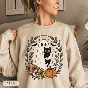 May include: A sand-colored crewneck sweatshirt featuring a graphic print of a white ghost holding a black cat, surrounded by a wreath of green leaves, orange and pink flowers, a pumpkin, and small bats. The text SAND is visible in a white box.