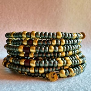 Könnte beinhalten: Ein mehrsträngiges Wickelarmband aus kleinen, runden Perlen in Metallic-Gold, mattem Dunkelgrün und Dunkelbraun. Das Armband ist mit einem kleinen, runden, goldfarbenen Metallanhänger versehen, in den ein Logo eingraviert ist.