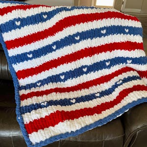 May include: A chunky knit blanket featuring horizontal stripes in red, white, and blue. The blue stripes are decorated with a repeating pattern of small white hearts. The blanket has a thick, textured yarn construction with a soft, plush appearance.