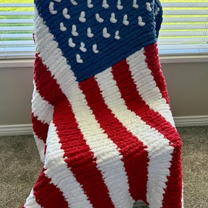 May include: A handmade, chunky knit blanket draped over a chair. The blanket features a design resembling the American flag, with red and white stripes and a blue field with white stars. The blanket is made of a soft, plush material.