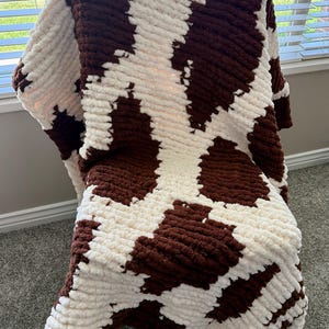 May include: A cozy, handmade blanket with a cow print design. The blanket features a pattern of large, irregular brown spots against a cream-colored background. The thick, plush material suggests warmth and comfort, perfect for home decor.