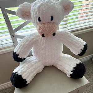 May include: A plush stuffed animal shaped like a cow, crafted from white, soft, textured yarn. The cow features a light pink snout, black embroidered eyes and nostrils, and black yarn accents on the tips of its four limbs and inner ears.