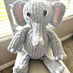 Pode incluir: Um peluche de elefante feito à mão em croché com fio grosso de cor cinzento-claro. O brinquedo apresenta orelhas grandes com detalhes interiores em rosa pálido, uma tromba comprida, olhos de segurança pretos e detalhes brancos nos pés.