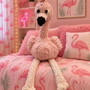 May include: A plush, hand-crocheted flamingo toy made from soft, light pink yarn. The flamingo features a long neck, a white face patch, a black beak, and large black and white eyes. It has two long, light-colored legs and a textured, fuzzy body.