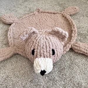 K&ouml;nnte beinhalten: Ein handgestrickter, b&auml;renf&ouml;rmiger Teppich aus weichem, grobem hellbraunem Garn. Der Teppich hat einen runden K&ouml;rper, vier ausgestreckte Gliedma&szlig;en, zwei abgerundete Ohren und eine markante wei&szlig;e Schnauze mit einer kleinen schwarzen Nase und zwei schwarzen ovalen Augen.