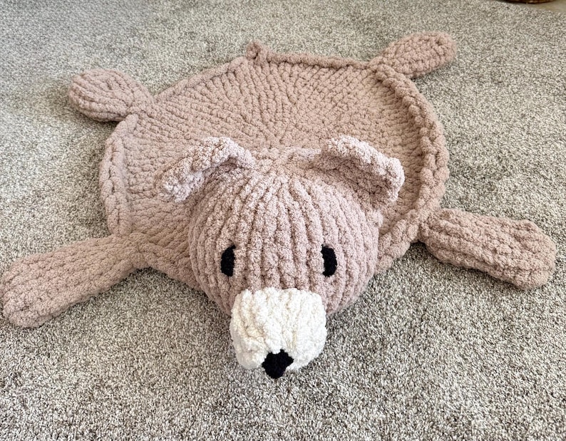 K&ouml;nnte beinhalten: Ein handgestrickter, b&auml;renf&ouml;rmiger Teppich aus weichem, grobem hellbraunem Garn. Der Teppich hat einen runden K&ouml;rper, vier ausgestreckte Gliedma&szlig;en, zwei abgerundete Ohren und eine markante wei&szlig;e Schnauze mit einer kleinen schwarzen Nase und zwei schwarzen ovalen Augen.