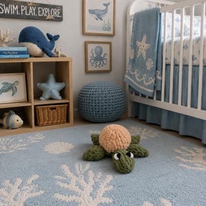 May include: A light blue area rug featuring a white coral and seashell pattern, with a plush, crocheted sea turtle toy in the center. The turtle has a tan shell, dark green limbs, and black and white eyes, resting on the textured, soft-pile rug surface.