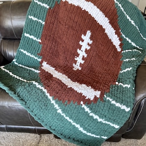 May include: A handmade, chunky knit blanket featuring a football design. The blanket has a brown football shape with white stripes and stitching, set against a green background with white stripes. The blanket is draped over a brown leather couch.