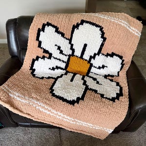 May include: A chunky knit blanket featuring a large, central daisy motif with white petals, a mustard yellow center, and black outlines. The background is a soft peach color, accented by horizontal white stripes near the top and bottom edges.