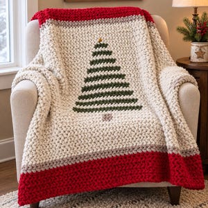 May include: A chunky knit throw blanket featuring a cream-colored base with a large green Christmas tree design in the center. The blanket has thick red and light gray horizontal stripes at the top and bottom edges, crafted from soft yarn.