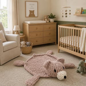 May include: A chunky, hand-knitted, dusty pink bear-shaped rug made of soft yarn. The rug features a round body, four limbs, and a detailed bear head with a cream-colored snout, black eyes, and a black nose, laid flat on a neutral-colored carpet.