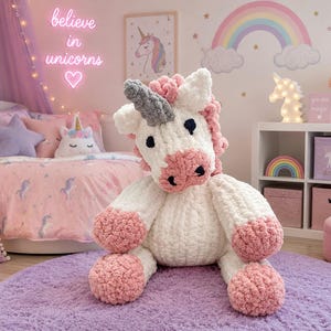 Puede incluir: Un peluche de unicornio tejido a mano con cuerpo blanco, pezuñas rosas, crin rosa y un cuerno gris texturizado. El animal de peluche presenta ojos y fosas nasales bordados en negro, confeccionado con hilo grueso y suave.