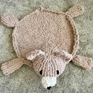 K&ouml;nnte beinhalten: Ein runder, hellbrauner, grob gestrickter Teppich in B&auml;renform. Der Teppich besteht aus einem runden K&ouml;rper, vier ausgestreckten Gliedma&szlig;en und einem B&auml;renkopf mit zwei Ohren, zwei schwarzen Augen und einer wei&szlig;en Schnauze mit kleiner schwarzer Nase.