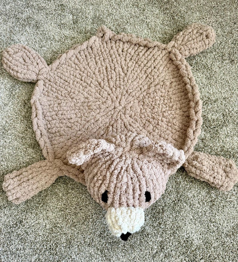 K&ouml;nnte beinhalten: Ein runder, hellbrauner, grob gestrickter Teppich in B&auml;renform. Der Teppich besteht aus einem runden K&ouml;rper, vier ausgestreckten Gliedma&szlig;en und einem B&auml;renkopf mit zwei Ohren, zwei schwarzen Augen und einer wei&szlig;en Schnauze mit kleiner schwarzer Nase.