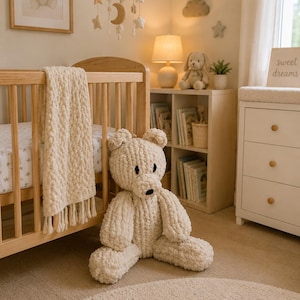 May include: A large, cream-colored, chunky knit plush bear sits on the floor next to a light wood crib. The crib features a matching cream-colored, fringed, chunky knit blanket draped over the side. The bear has black embroidered eyes and nose.