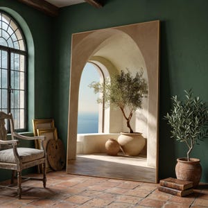 May include: A large, rectangular mirror with a light brown frame. The mirror reflects an arched doorway with a potted olive tree, a view of the ocean, and a terracotta tiled floor. The room has green walls and a wooden chair.