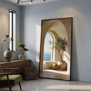 May include: A large, rectangular mirror with a black frame leans against a grey wall. The mirror reflects an outdoor scene with an olive tree in a pot, an arched doorway, and a view of the sea. The room features a green chair and wooden furniture.