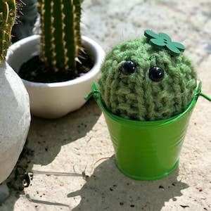 Op de afbeelding: Een handgemaakte groene gebreide cactus met grote zwarte ogen en een houten klaver, in een kleine groene metalen emmer. Andere cactussen in witte potten staan op de achtergrond.