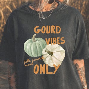 May include: A charcoal gray short-sleeve t-shirt featuring the orange text GOURD VIBES ONLY and the smaller text falls favorite mood. The graphic includes a light green pumpkin and a white pumpkin centered on the front of the garment.