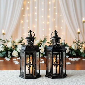 May include: Two black metal lanterns with glass panels, each with a candle inside. The lanterns have ornate detailing and a handle on top. They are set on a white runner, with white flowers and a curtain backdrop.