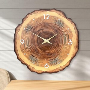 May include: A round wooden wall clock designed to resemble a cross-section of a tree trunk. The clock face features a natural wood grain pattern with white numerals at the 12, 3, 6, and 9 o'clock positions. The clock hands are gold-colored.