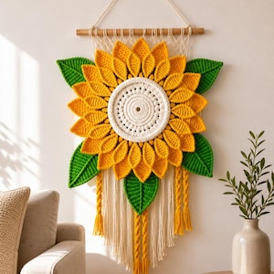 May include: A crochet sunflower wall hanging with yellow petals, a cream centre, and green leaves. It's suspended from a wooden dowel with cream fringe and yellow braided details. This handmade piece brings a touch of nature to interior decor, measuring approximately 50cm in diameter.