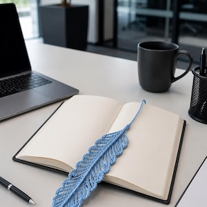 May include: A light blue, crocheted feather bookmark rests on an open notebook. The bookmark is made of yarn and has a detailed, textured design. A black pen, a black mug, and a laptop are also on the desk.