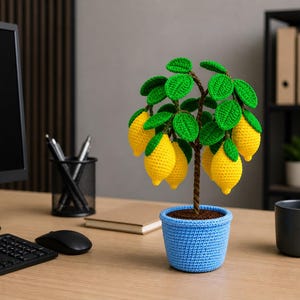 May include: A crocheted lemon tree in a blue pot sits on a wooden desk. The tree features bright yellow lemons, green leaves, and a brown trunk. A computer monitor, keyboard, and mouse are in the background.