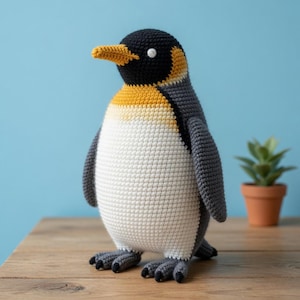 Può includere: Un pinguino all'uncinetto con testa nera, pancia bianca e ali grigie. Il pinguino presenta un becco e una macchia sul collo di colore giallo-arancio, con piccole zampe nere. La texture è composta da punti di filato visibili sul corpo.