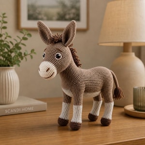 May include: A crocheted donkey figurine in shades of brown and white. The donkey has large ears, a fluffy mane, and a small tail. The figurine is standing on a wooden surface.