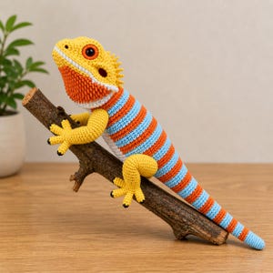 Könnte beinhalten: Eine gehäkelte Eidechse in gelben, orangefarbenen und blauen Streifen sitzt auf einem braunen Ast. Die Eidechse hat einen gelben Kopf mit orangefarbenen und weißen Akzenten und gelbe Füße mit schwarzen Krallen. Der Schwanz ist lang und gestreift.