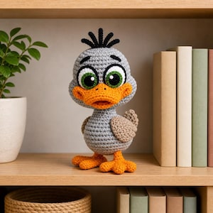 Op de afbeelding: Een handgemaakte grijze gehaakte eend met een oranje snavel en poten, en grote groene en witte ogen. De eend heeft een kleine pluk zwart garen op zijn hoofd. Het staat op een houten plank met boeken en een plant.