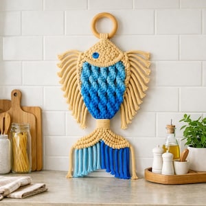May include: A macrame fish wall hanging with a wooden ring at the top. The fish features a blue and beige color scheme, with a blue gradient body and beige fins and tail. A blue bead serves as the eye.