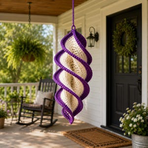 May include: A hanging, spiral-shaped crochet decoration with a cream-colored center and a vibrant purple border. The decorative piece is suspended from a purple cord and hangs on a porch. A rocking chair and hanging fern are visible in the background.