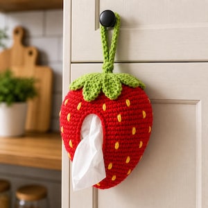 Peut inclure: Un porte-mouchoirs en forme de fraise rouge crocheté, avec des détails de graines jaunes et un dessus vert en forme de feuilles. Le support est suspendu à un bouton noir sur une porte d'armoire blanche. Des mouchoirs blancs sont visibles.