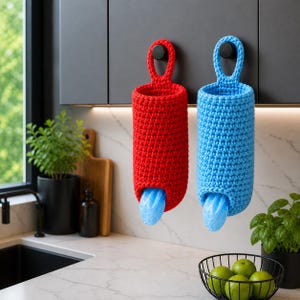 May include: Two crocheted bag dispensers, one red and one blue, hang on a kitchen cabinet. Each dispenser has a loop for hanging and a bottom opening with a blue plastic bag visible. The dispensers are cylindrical.
