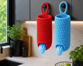 Op de afbeelding: Twee gehaakte zakdispensers, één rood en één blauw, hangen aan een keukenkast. Elke dispenser heeft een lus om op te hangen en een opening aan de onderkant met een blauwe plastic zak zichtbaar. De dispensers zijn cilindrisch.