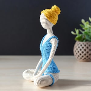Puede incluir: Muñeca de ganchillo hecha a mano en postura de yoga sentada. La muñeca tiene un cuerpo color crema, un traje de baño azul y un moño amarillo. La muñeca está sentada con las piernas cruzadas y los brazos apoyados en las piernas. El fondo es gris oscuro.