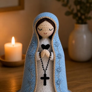 May include: A handmade crochet doll depicting the Virgin Mary. The doll wears a light blue hooded cloak with floral accents, a white dress, and a black rosary with a cross. The doll's face has closed eyes and a gentle smile.
