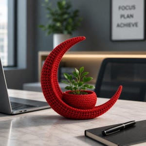 May include: A red, crescent-shaped crochet planter holding a small green plant. The planter is on a light-colored desk with a laptop and a notebook. The words "FOCUS PLAN ACHIEVE" are visible in the background.