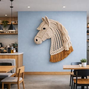 May include: A large, beige macrame horse head wall hanging with a tan mane and fringe. The horse head is mounted on a light blue wall in a cafe setting, with wooden tables and chairs visible in the foreground.