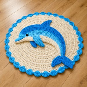 May include: A handmade, round crochet rug featuring a blue dolphin design. The dolphin has varying shades of blue and white, with a black eye. The rug has a cream-colored center and a scalloped blue border. The rug is placed on a wooden floor.