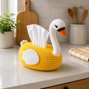 Puede incluir: Una funda de caja de pañuelos de ganchillo hecha a mano con forma de cisne. El cisne tiene un cuerpo amarillo, alas blancas y cuello y cabeza blancos. El pico del cisne es naranja y los ojos son negros. Se ven pañuelos blancos.
